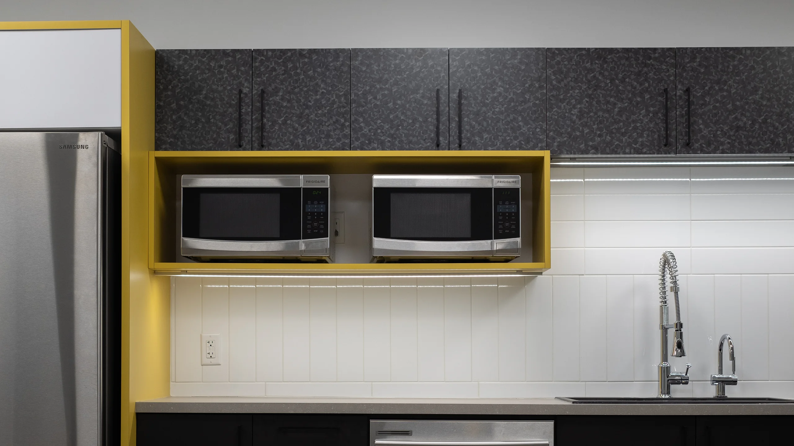 Office kitchen with bright yellow accent cabinetry framing stainless steel refrigerator and dual microwave cubby